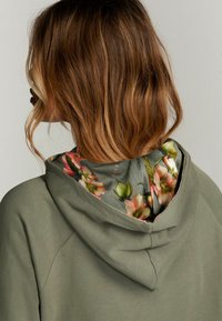 Groene hoodie met een bloemenpatroon aan de voering van de capuchon. Gladde textuur, raglanmouwen en een relaxte pasvorm. Achteraanzicht van het kledingstuk.