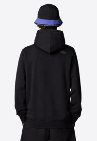 Black hoodie with a large hood and ribbed cuffs, featuring a small green logo on the back. Paired with a black and blue bucket hat.