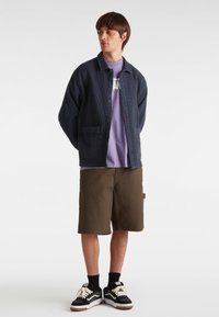 Giacca blu navy con bottoni in tessuto testurizzato sopra una maglietta grafica lilla, abbinata a pantaloni corti marroni e sneakers nere con dettagli bianchi.