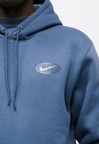 Μπλε φούτερ με κουκούλα και κορδόνια, που έχει μικρό λευκό λογότυπο γήινου σχήματος με ραβδώσεις στο στήθος, φορεμένο από άτομο με σκούρο δέρμα.