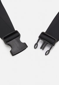 Sangle en nylon noir avec des boucles en plastique. Une boucle présente deux pointes, tandis que l'autre a un design à glissière et crochet. La texture est lisse.