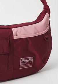 Bordeauxrood en lichtroze Columbia schoudertas met voorpatchlogo, ritszak en verstelbare schouderband tegen een witte achtergrond.