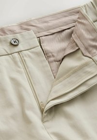 Pantaloni beige con chiusura a pulsante, apertura con zip e una fodera liscia di un rosa chiaro. Cuciture visibili e dettagli della trama del tessuto.