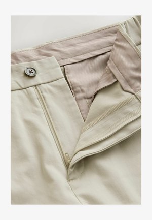 Beige Hosen mit einem Knopfverschluss, Reißverschluss und einem glatten, hellroten Innenfutter. Sichtbare Nähte und Stofftexturdetaillierung.