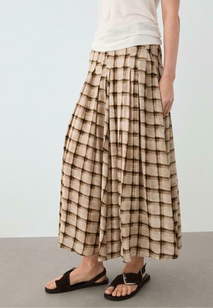 Personne portant un pantalon large à carreaux beige et marron avec un design plissé, un haut blanc et des sandales noires à boucle.