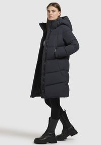 Lang marineblå pufferjakke med hætte. Har lynlås foran, store lommer og quiltet design. Båret over sorte leggings og snørelæderstøvler.