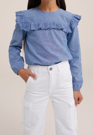 Fille portant un haut bleu à manches longues avec des volants en dentelle et un pantalon cargo blanc, une main dans la poche, vue du cou aux hanches.