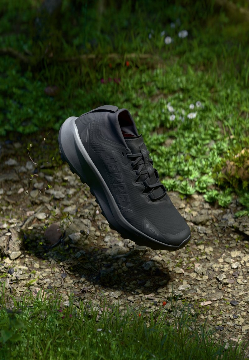 Chaussure de sport noire avec une tige texturée, un système de laçage et une semelle gris clair, survolant une surface rocheuse et herbeuse.