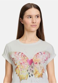 Lichtgrijze t-shirt met een grote vlinderafbeelding in pastelkleuren met een roze hart, gedetailleerd met bloemaccenten en sprankels.