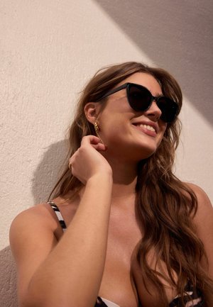 Woman wearing black cat-eye sunglasses and gold hoop earrings, smiling with hand near her neck against a light textured wall.