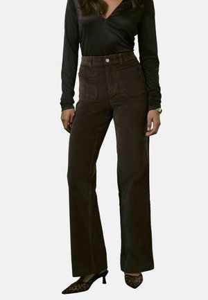 Braune Cord-Hosen mit hohem Bund und weitem Bein, mit aufgesetzten Taschen und Knopfverschluss. Kombiniert mit einem schwarzen Langarmoberteil.
