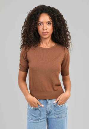 Femme aux cheveux bouclés portant un haut marron à manches courtes et un jean bleu avec des détails tressés sur les poches, debout les mains dans les poches.
