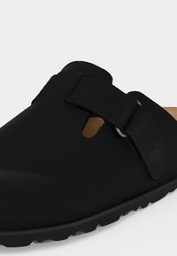 Μαύρο παπούτσι τύπου slip-on με ρυθμιζόμενο λουράκι και μεταλλικό πόμολο, με διαμορφωμένη σόλα και ανοιχτή πλευρική λεπτομέρεια.
