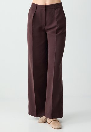 Szerokie spodnie o ciemnym brązowym kolorze z przodu z zakładkami, szlufkami na pasek oraz beżowe wsuwane buty na osobie stojącej na jednolitym tle.