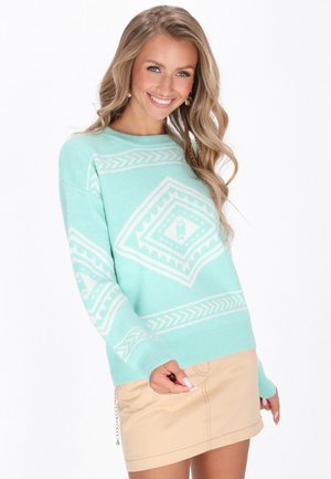 Jonge vrouw glimlacht, draagt een muntgroene trui met witte geometrische patronen en een beige rok, staat tegen een witte achtergrond.