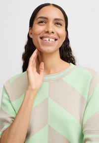 Hellgrüner und beiger gemusterter Pullover mit einem Zickzackdesign, rundem Ausschnitt und kurzen Ärmeln, getragen von einer lächelnden Person.