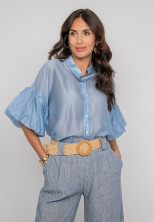 Femme portant un chemisier léger bleu clair transparent avec des manches bouffantes, une large ceinture et un pantalon bleu clair taille haute, posant les mains dans les poches.