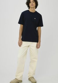 Jeune homme debout portant un t-shirt bleu marine avec un petit logo, un pantalon couleur crème et des chaussures beige sur un fond clair uni.