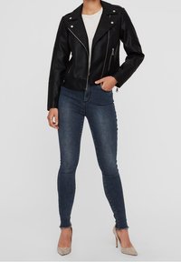 Giacca da motociclista nera in ecopelle con cerniere e borchie argento, indossata sopra una camicia bianca e abbinata a jeans scuri aderenti e tacchi beige.