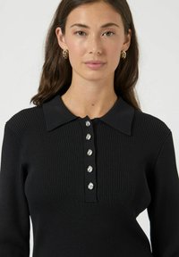 Top nero a maniche lunghe con coste, dotato di colletto abbottonato e sette bottoni metallici lungo il davanti.