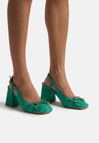 Tacchi slingback verde in suede con finitura texturizzata, punta quadrata e tacco spesso. Presentano un fiocco decorativo con dettagli in strass.