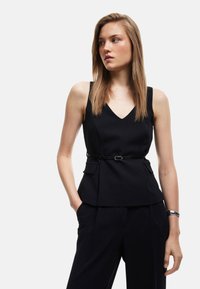 Sort ærmeløs top med V-hals, struktureret peplum-design, to sidelommer og et tyndt bælte i taljen. Båret sammen med matchende bukser.