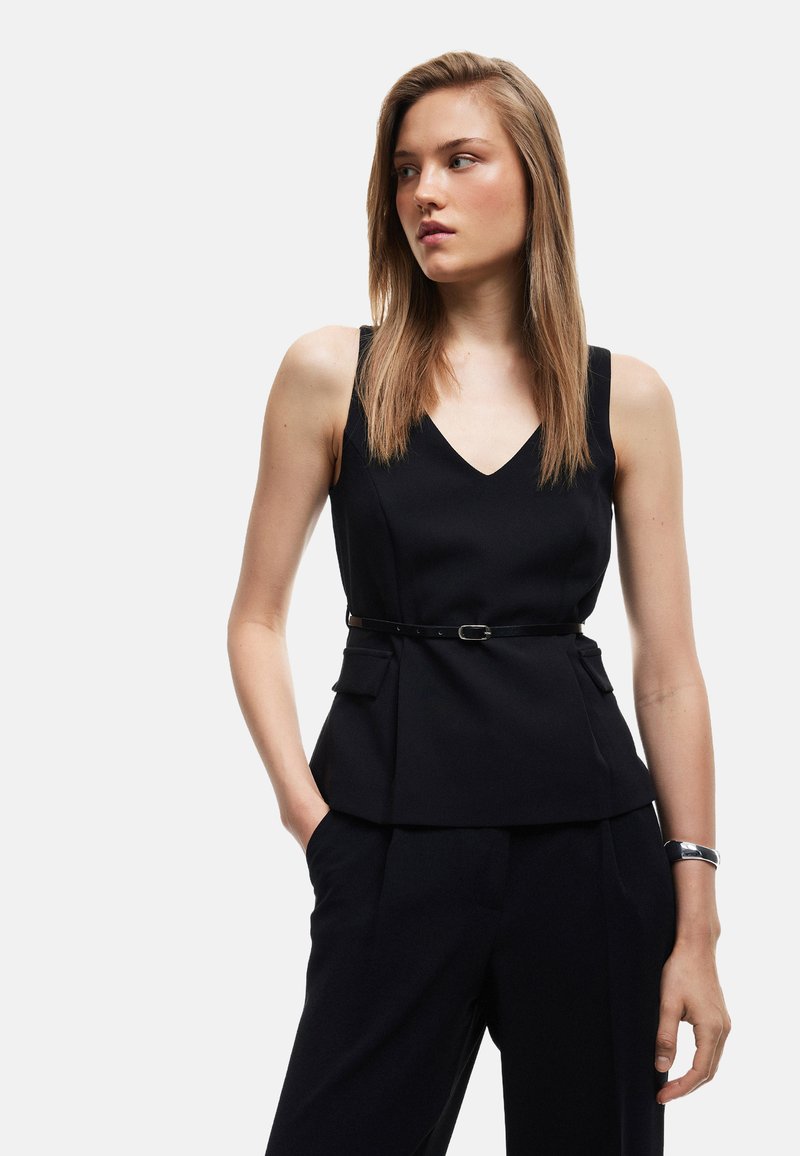 Sort ærmeløs top med V-hals, struktureret peplum-design, to sidelommer og et tyndt bælte i taljen. Båret sammen med matchende bukser.