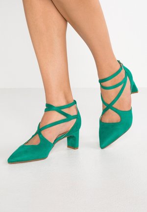 Pieds portant des chaussures en daim vert à talons moyens, bouts pointus et brides croisées à la cheville, sur un fond blanc.