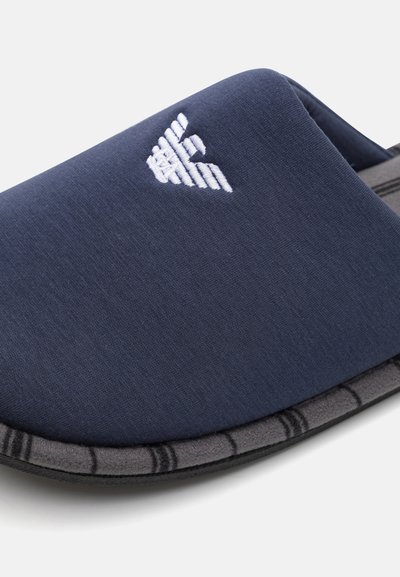 Pantofola grigia e blu navy senza lacci con tomaia in tessuto, caratterizzata da un logo ricamato bianco e una suola grigia testurizzata con un bordo decorato.