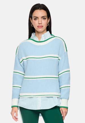 Frau mit glattem dunklem Haar trägt einen blau-weißen Strickpullover mit grünen Streifen über einem grün gestreiften Hemd mit Knöpfen und dunkelgrünen Hosen.