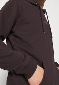 Ciemnobrązowa bluza z kapturem z regulowanym sznurkiem, przednią kieszenią typu kangurka, ściągaczami na rękawach oraz haftowanym logo na piersi. Miękka faktura materiału.