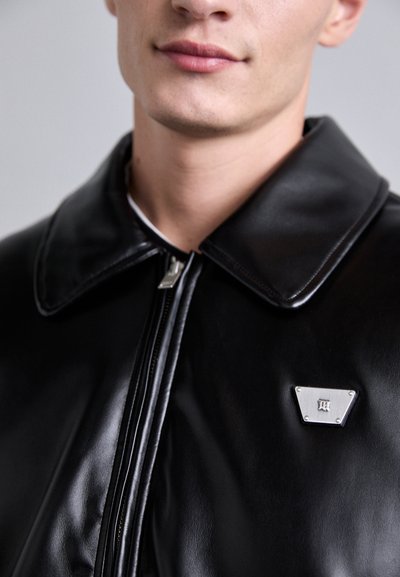 Jeune personne portant une veste en cuir noir avec une fermeture éclair à l'avant et une petite épingle triangulaire en argent sur la poitrine.