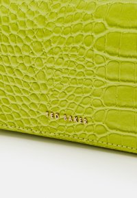 Portafoglio in pelle verde con impresso coccodrillo, dalla texture liscia e logo dorato "TED BAKER". Presenta bordi cuciti per dettagli aggiuntivi.