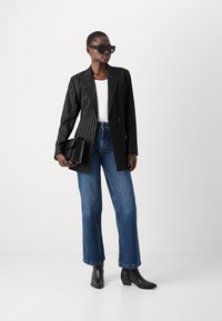 Svart pinstripet blazer med to knapper, hvit topp, høytlivde blå jeans og svarte ankelstøvler. Modellen holder en teksturert svart håndveske.