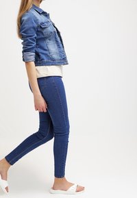 Žena jde, má na sobě shrnutou denimovou bundu, béžový top, modré skinny džíny a bílé pantofle na pozadí bílé barvy.