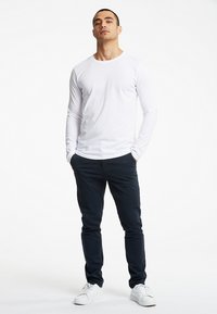 Hvid langærmet skjorte, navy bukser og hvide sneakers. Model står oprejst og fremviser et tætsiddende design og en glat tekstur.