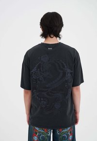 Czarny oversize t-shirt z ciemnym wzorem smoka na plecach. Ma okrągły dekolt i krótkie rękawy, a jego powierzchnia jest gładka.