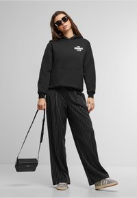 Black hoodie with "THE BRUNCH CLUB" text, paired with black pinstripe trousers. Accessories include black sunglasses and a small crossbody bag.