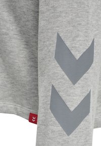 Chemise gris à manches longues en tissu doux, arborant un motif chevron gris sur la manche et une petite étiquette rouge en accent à l'ourlet.