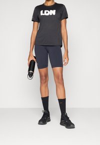 Camiseta atlética negra con gráfico "LDN", combinada con pantalones cortos oscuros, zapatillas deportivas negras y calcetines negros. Sostiene una botella de agua negra.