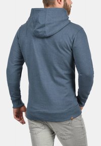 Blå hettegenser laget av myk stoff, med kengurulomme, ribbestrikkede mansjetter og hette med snor. Minimalistisk design uten fremtredende mønstre.