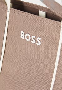 Canvas boodschappentas in taupe met witte "BOSS" logo. Heeft contrasterende witte handvatten en een structuurstof met zichtbare stiksels.