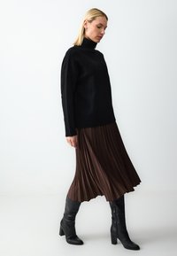 Maglione a collo alto nero con una vestibilità rilassata, abbinato a una gonna a pieghe marrone di lunghezza al ginocchio e stivaletti neri con tacco alti fino al ginocchio.