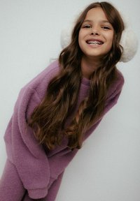 Fioletowy dziany sweter i dopasowane spodnie, długie brązowe włosy i białe puszyste nauszniki, uśmiechająca się na jednolitym tle.