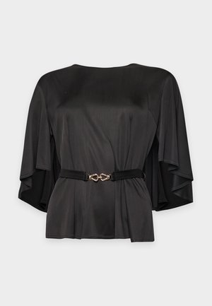 Une femme porte une robe midi noire à manches cape et une ceinture fine, assortie de chaussures à talons pointues en résille, sur un fond de studio uni.