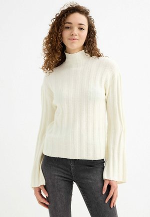 Suéter crema de canalé con cuello alto y mangas acampanadas. Textura suave y corte holgado, combinado con jeans gris oscuro.