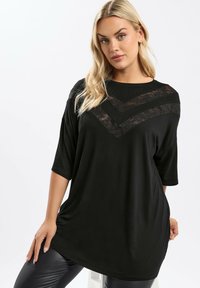 Blond kvinna i en lös svart topp med spetsshalsband och svarta läderbyxor, poserande mot en vit bakgrund.