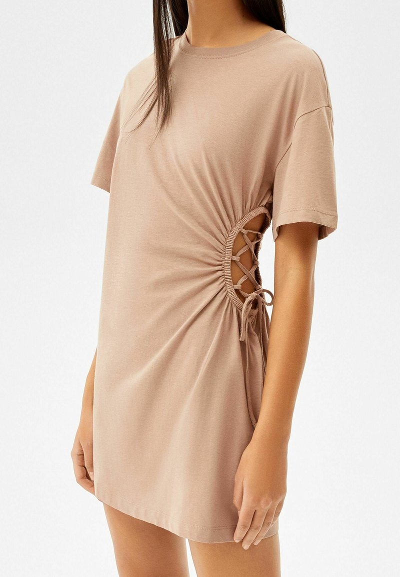 Femme portant une robe beige à manches courtes avec un côté froncé et un laçage laissant apparaître la peau à travers l'ouverture.