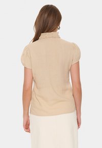  Béžová blúzka s volánikmi na golieri a krátkymi puff rukávmi; hladká textura s jemným leskom. Nosená s off-white sukňou.