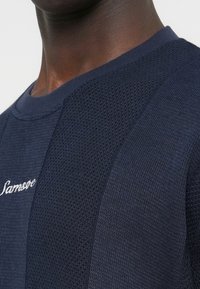 Gros plan sur une personne portant un pull en maille texturée bleu marine à col rond avec des lettres blanches brodées sur la poitrine.